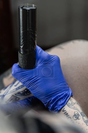 Foto vertical: un primer plano de un artista del tatuaje con guantes agarrando la máquina de tatuaje, preparada para crear arte permanente en la piel. Concepto negocio, arte.