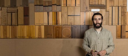 Photo for Horizontal photo a bearded woodworker mid adult caucasian stands confidently in his workshop, a wall of labeled wood samples showcasing various species behind him. Copy space. Business concept. - Royalty Free Image