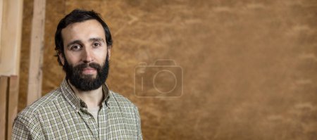 Horizontal photo a serene portrait of a bearded craftsman mid adult caucasian in a plaid shirt, with a soft smile and a textured wooden wall in the background. Copy space. Business concept.