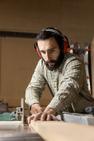 Vertical photo Captured from a dynamic angle, a man mid adult carpenter with ear protection is fully absorbed in operating a table saw, ensuring each cut is made with precision. Business concept.