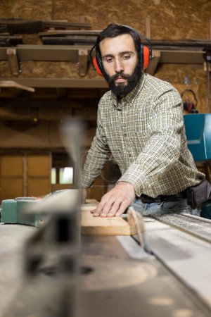 Vertical photo focused carpenter with hearing protection uses a table saw to cut a wooden board in a well-equipped woodworking workshop. Business concept.