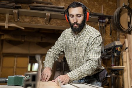 Horizontal photo serious carpenter with hearing protection operates a table saw, pushing a wooden board towards the rotating blade in a busy woodshop. Business concept.