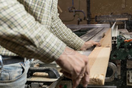 Horizontale Foto-Nahaufnahme der Hände eines Tischlers, wie er ein Stück Holz manuell in eine Tischkreissäge einfüttert, während in der Werkstatt Sägemehl wirbelt. Geschäftskonzept.