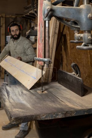 Vertical photo a man mid adult caucasian carpenter with protective ear gear operates a bandsaw, cutting a wooden plank with precision in a dusty workshop environment. Business concept.