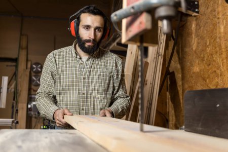 Photo for Horizontal photo an attentive carpenter man mid adult caucasian with protective ear muffs guides a wooden board through a bandsaw, demonstrating precision in his craft. Business concept. - Royalty Free Image