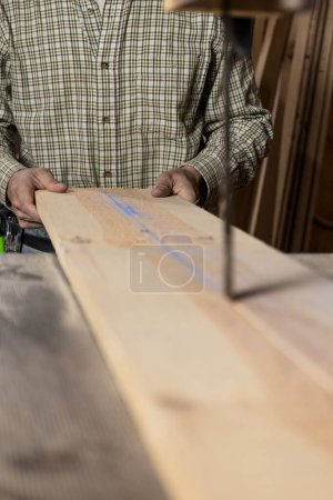 Vertikale Fotohände eines Zimmermanns, Mitte erwachsener Kaukasier, richten ein Holzbrett vorsichtig zu einem Bandsägeblatt aus, wobei der Schwerpunkt auf der markierten Richtlinie für einen präzisen Schnitt liegt. Kopierraum. Geschäftskonzept.
