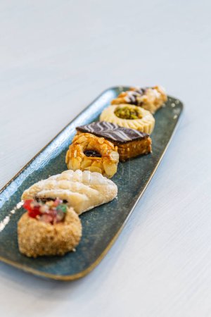 Foto vertical: una selección seleccionada de pasteles árabes, cada uno con una cubierta y un diseño únicos, servidos en una bandeja de cerámica azul elegante sobre un fondo neutro. Concepto de alimentación y cultura.