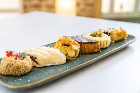 Horizontales Foto einer eleganten Ausbreitung gemischter arabischer Süßigkeiten, mit vielfältigen Aromen und Texturen, kunstvoll arrangiert auf einem strukturierten blauen Servierteller. Ernährungs- und Kulturkonzept.