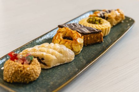 Horizontal photo a tasteful arrangement of Arabic pastry delicacies with various toppings, artistically served on a textured blue platter, ideal for high tea or a dessert buffet. Food and culture concept.