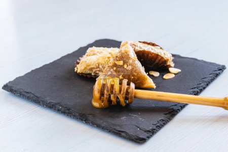 Horizontales Foto einer Auswahl honigglasierter arabischer Süßigkeiten mit Nüssen, elegant serviert auf einer dunklen Steinplatte, wobei ein hölzerner Honiglöffel dem Ganzen eine natürliche Note verleiht. Ernährungs- und Kulturkonzept.