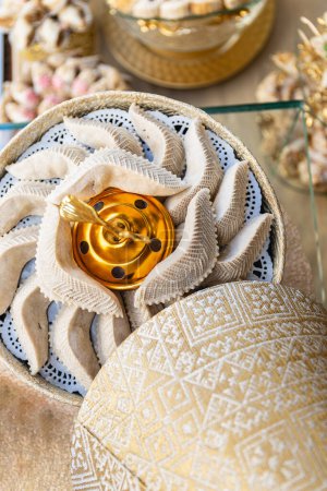 Vertikales Foto eines künstlerischen goldenen Mittelstücks, umgeben von halbmondförmigen arabischen Süßigkeiten, präsentiert auf einem Tisch mit zarten Spitzendetails. Ernährungs- und Kulturkonzept.