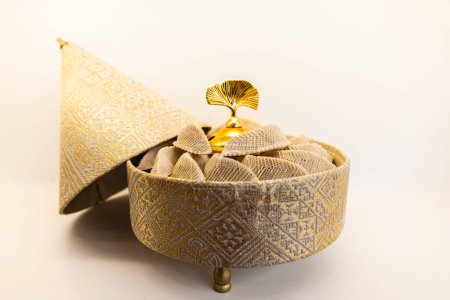 Horizontales Foto Ein goldener, bestickter Behälter, kunstvoll mit einem geometrischen Muster gestaltet, hält eine Auswahl traditioneller arabischer Backwaren vor einem minimalistischen Hintergrund. Ernährungs- und Kulturkonzept.