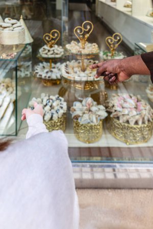 Vertical photo a close-up hand in hand with two people pointing to an array of finely decorated Arabic sweets displayed in a shop window, showcasing the rich variety. Food and culture concept.