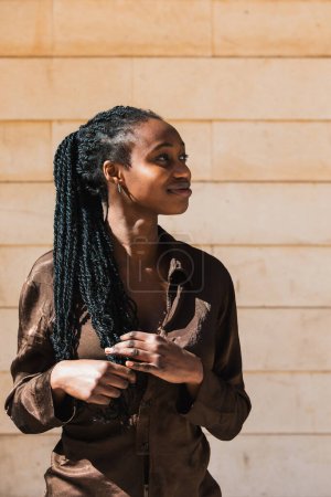 Vertical photo sunlit portrait of a young woman african with elegant braids, looking aside with a subtle smile, against a warm stone wall background. People concept.