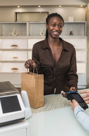 Vertical photo a delighted customer holds a paper bag of pastries, completing a purchase with a contactless payment at a modern bakery's checkout counter. Food and culture concept.