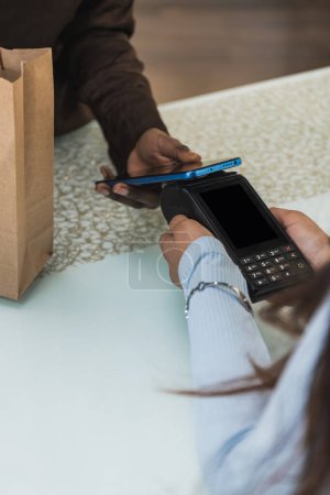 Vertical photo a close-up image captures the moment of a contactless payment, showing a customer's hand holding a smartphone over a card reader at a bakery. Business concept.