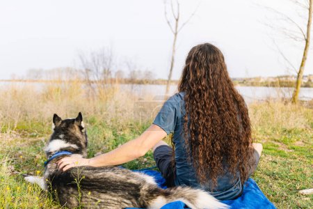 Horizontales Foto eines Mannes mit wallenden lockigen Haaren, der sich am Ufer des Flusses entspannt und einen ruhigen Moment mit seinem treuen Husky in der sanften Umarmung der Natur genießt. Lifestyle-Konzept.