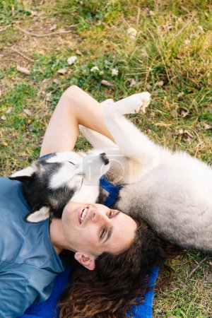Vertical photo laughter fills the air as a man with curly hair and his playful husky share a moment of pure bliss, lying on the grass together. Lifestyle concept.