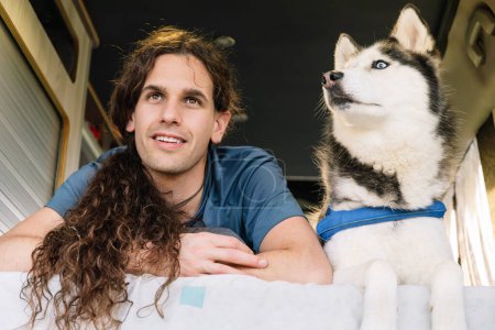 Horizontal photo a relaxed man with curly hair and his blue-eyed husky comfortably lounge inside a camper van, enjoying a moment's rest during their travels. Lifestyle concept.