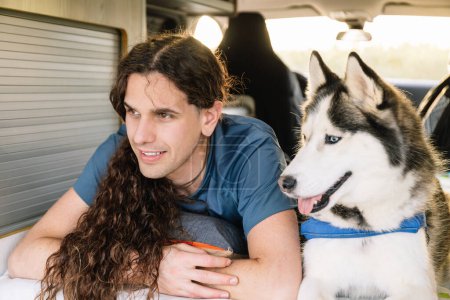 Horizontal photo a man with luscious curly hair shares a relaxed moment with his joyful husky inside the comfortable space of their modern camper van. Lifestyle concept.