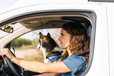 Horizontal photo young man with long curly hair focusing on the road as he drives his camper van, with his husky companion enjoying the view from the passenger seat. Lifestyle concept.