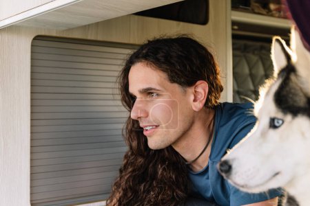 Horizontal photo a smiling man with curly hair and his attentive husky companion look out together from the comfort of their camper van, capturing a moment of shared adventure. Lifestyle.