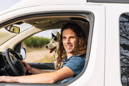 Horizontal photo a smiling man with long curly locks shares the driver's seat with his spirited husky, both radiating happiness on their camper van escapade. Lifestyle concept.