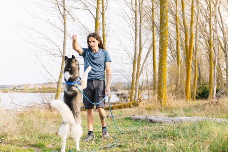 Horizontal photo active young man with long hair engages his attentive husky in a training exercise amid the golden trees of a serene riverside landscape. Lifestyle concept.