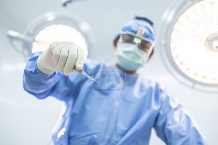 Photo for Uprisen of surgeon or doctor holding scalpel inside operating room in hospital with blurred surgical lamp background.Surgeon in blue uniform did surgery technology.Selective focus at hand of people. - Royalty Free Image