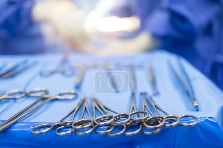 Ein medizinisches oder chirurgisches Instrument für die Operation im Operationssaal mit einem verschwommenen Team von Ärzten in blauem Kittelhintergrund. Chirurgische Klammern für einen Chirurgen auf dem blauen Tablett. Hintergrund mit Licht verschwimmen.