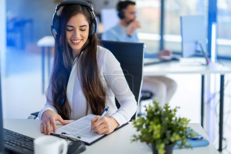 Photo for Dedicated operator woman wearing headset, skillfully multi-tasking by taking notes and actively listening with to Clients needs in a Call Center office - Royalty Free Image