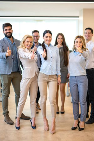 Full length of large group of confident business people showing thumbs up standing close to each other and smiling at office. Successful business team