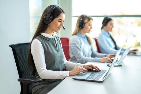 Téléchargez les photos : Groupe de trois femmes opératrices de soutien à la clientèle assises en rangée et travaillant sur des ordinateurs portables au bureau du centre d'appels - en image libre de droit