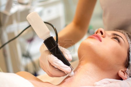 Beauty doctor with ultrasonic scraber doing procedure of ultrasonic cleaning of female clients neck.
