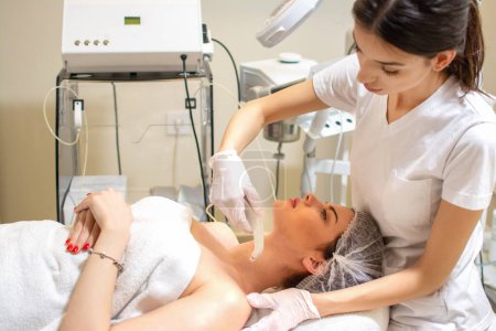 Téléchargez les photos : Esthéticienne appliquant un traitement de la peau avec un appareil moderne à la jeune femme au salon de spa - en image libre de droit