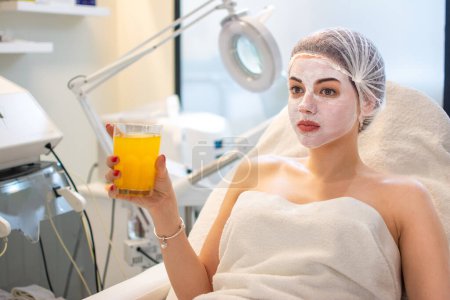 Téléchargez les photos : Belle femme ayant un traitement du visage et boire du jus d'orange au salon de beauté. - en image libre de droit