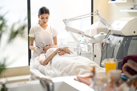 Téléchargez les photos : Esthéticienne faisant des procédures de cosmétologie en utilisant une machine de beauté à radiofréquence pour les soins de la peau dans un salon de beauté moderne - en image libre de droit