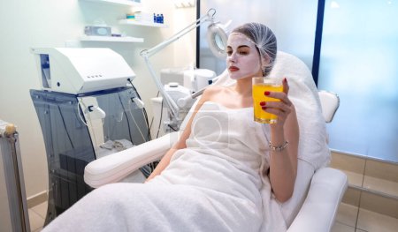 Téléchargez les photos : Belle femme avec masque en argile buvant du jus d'orange à la clinique de beauté - en image libre de droit