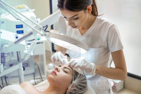 Téléchargez les photos : Esthéticienne serrant l'acné et les boutons du nez des jeunes femmes à l'aide d'une lampe et d'une loupe dans une clinique de spa de beauté - en image libre de droit