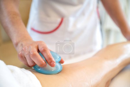 Téléchargez les photos : Gros plan de la main féminine tenant champignon en forme Gua Sha anti outil de cellulite et faire massage sur les jambes des clients dans le salon de beauté. - en image libre de droit