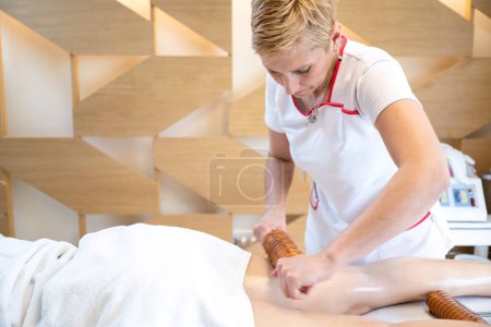 Photo for Therapist using wooden massage roller in maderotherapy for cellulite reduction on female client legs at beauty salon - Royalty Free Image