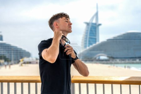 Joven guapo que se pone gafas de sol alrededor de la camisa del cuello mientras está de pie en la playa de Dubai