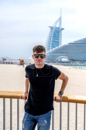 Porträt eines jungen Mannes in lässiger Sommerkleidung mit Sonnenbrille, der am Zaun lehnt und am Strand von Dubai posiert