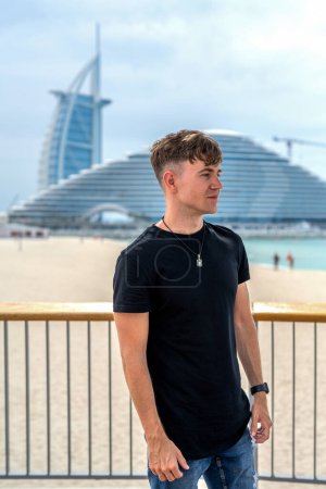 Hombre joven guapo en ropa casual de pie cerca de la cerca y mirando por encima de la playa en Dubai