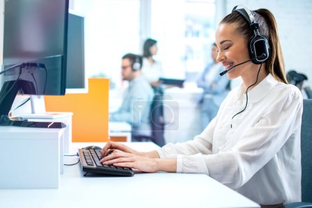 Photo for Dedicated smiling woman in headset using computer in office. Call center, customer service support concept. - Royalty Free Image