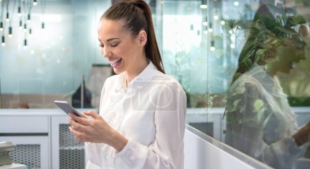Photo for Smiling happy woman in formal shirt with tied hair texting messages on her smart phone at office. - Royalty Free Image