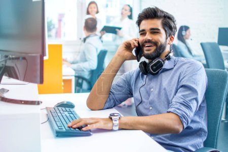 Photo for Smiling handsome middle eastern of south asian ethnicity man employee in business wear talking on mobile phone at his workspace at computer in office. - Royalty Free Image