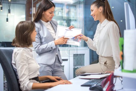 Photo for Three business women discussing about company financial growth in past months pointing over graphs on paper document during informal meeting at office. - Royalty Free Image