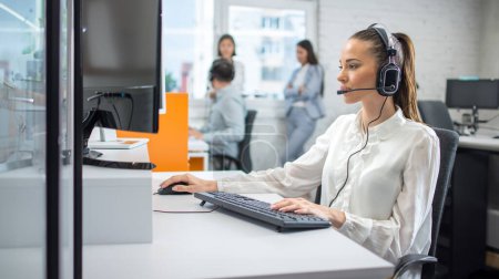 Téléchargez les photos : Jolie jeune femme avec casque de travail sur ordinateur tout en parlant avec le client dans le centre d'appels. Assistance clientèle en ligne. - en image libre de droit