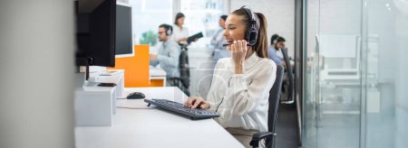 Téléchargez les photos : Femme d'affaires souriante utilisant un ordinateur et écoutant le client via un casque dans le bureau du centre d'appels. Vue panoramique. - en image libre de droit